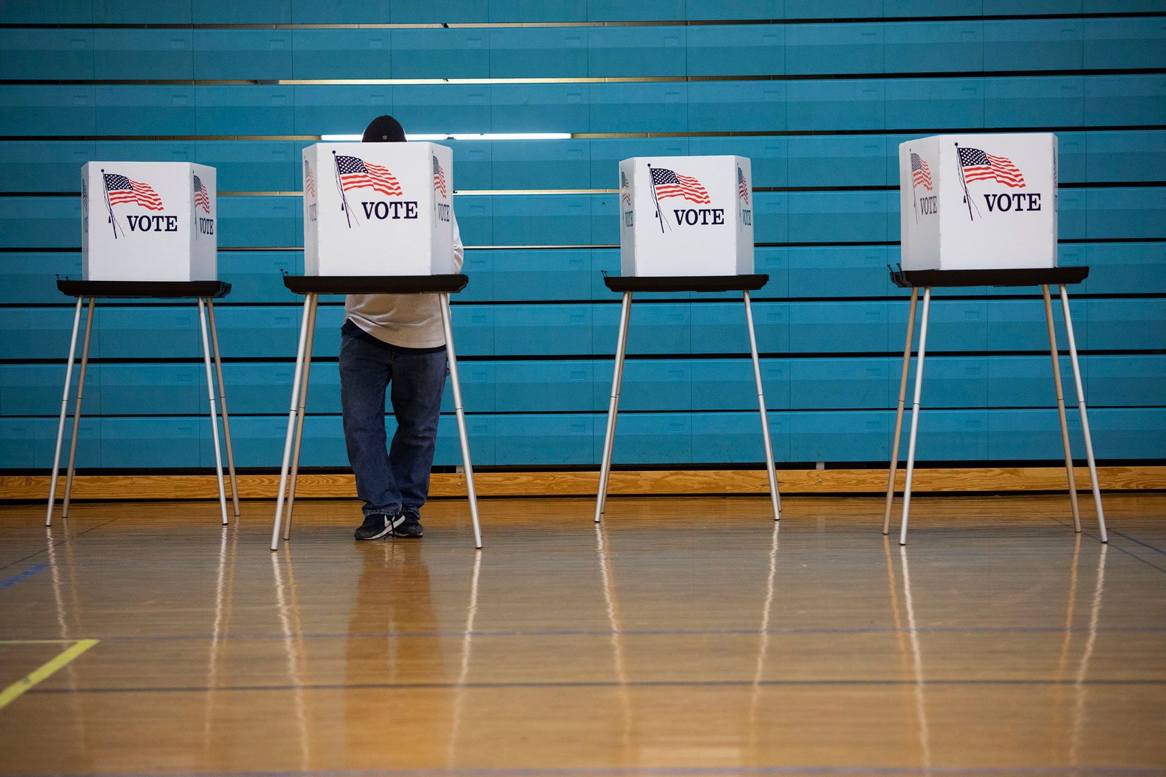 Four voting booths are seen in front of a blue wall, and a voter fills out their ballot in one of the booths.