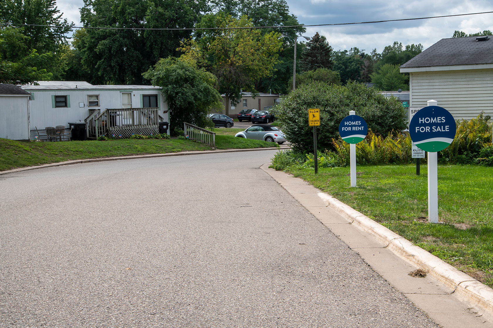 Photo shows a lawn with signs advertising homes for rent and for sale, with mobile homes in the background