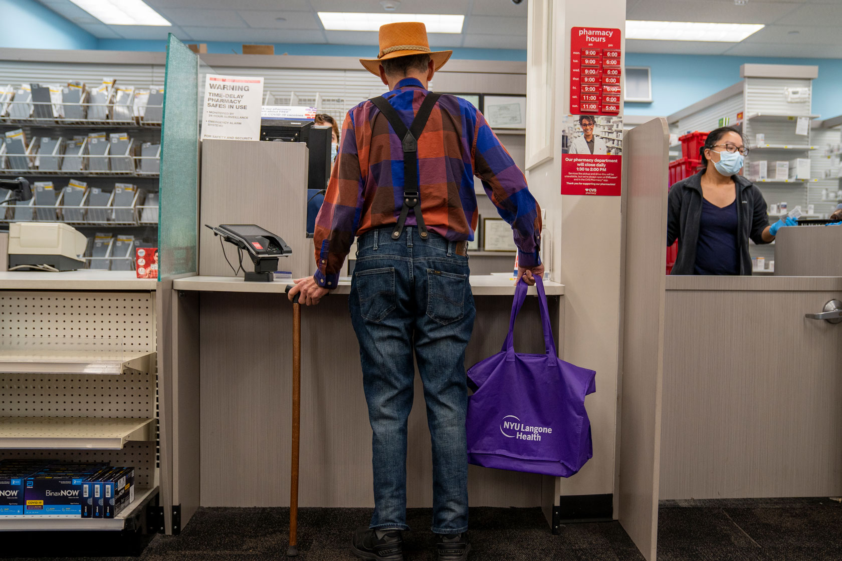 The back of a man holding a cane in his left hand is seen at a pharmacy counter with a pharmacy worker in the background.