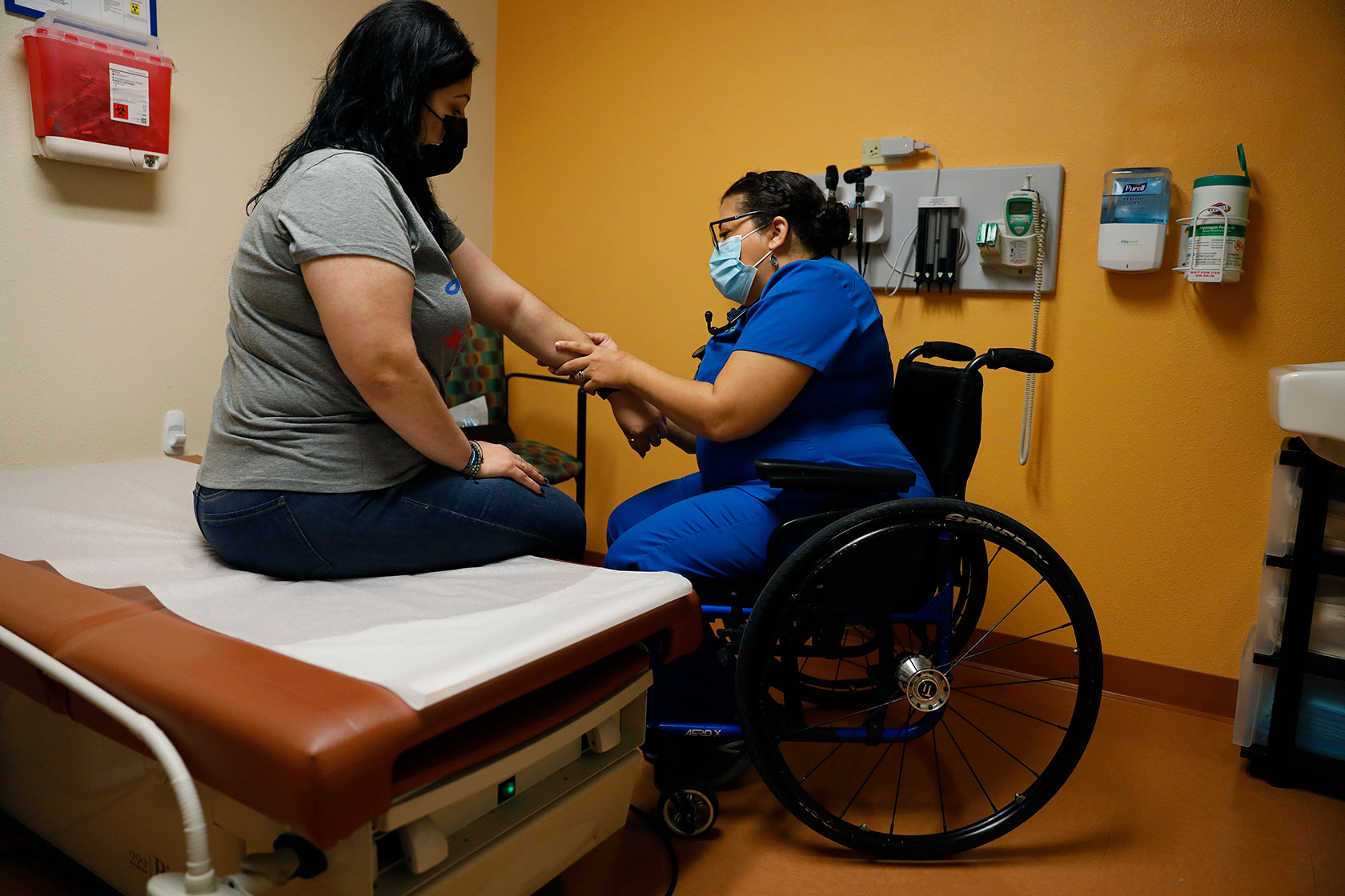 Photo shows a woman sitting on an exam table, with a doctor in blue scrubs seated in a wheelchair holding the patient's arm, in a yellow painted room