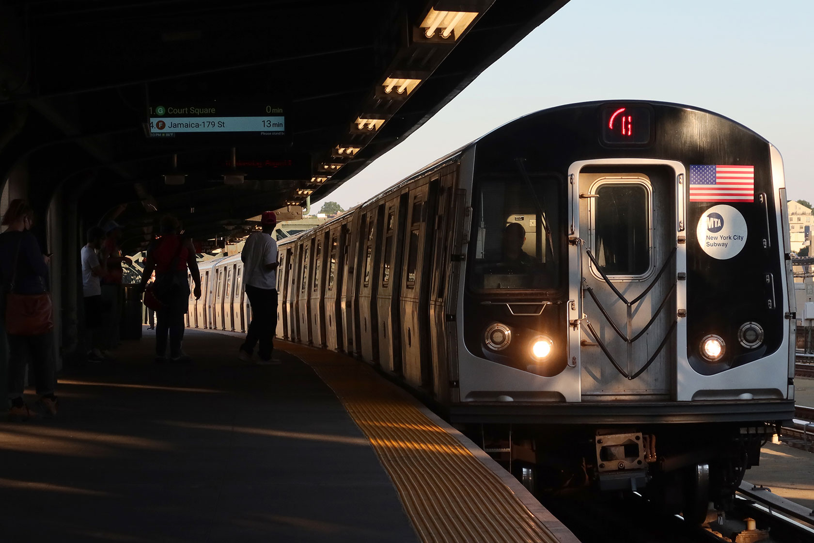 Photo shows a train pulling up to a covered platform with two people standing near the edge