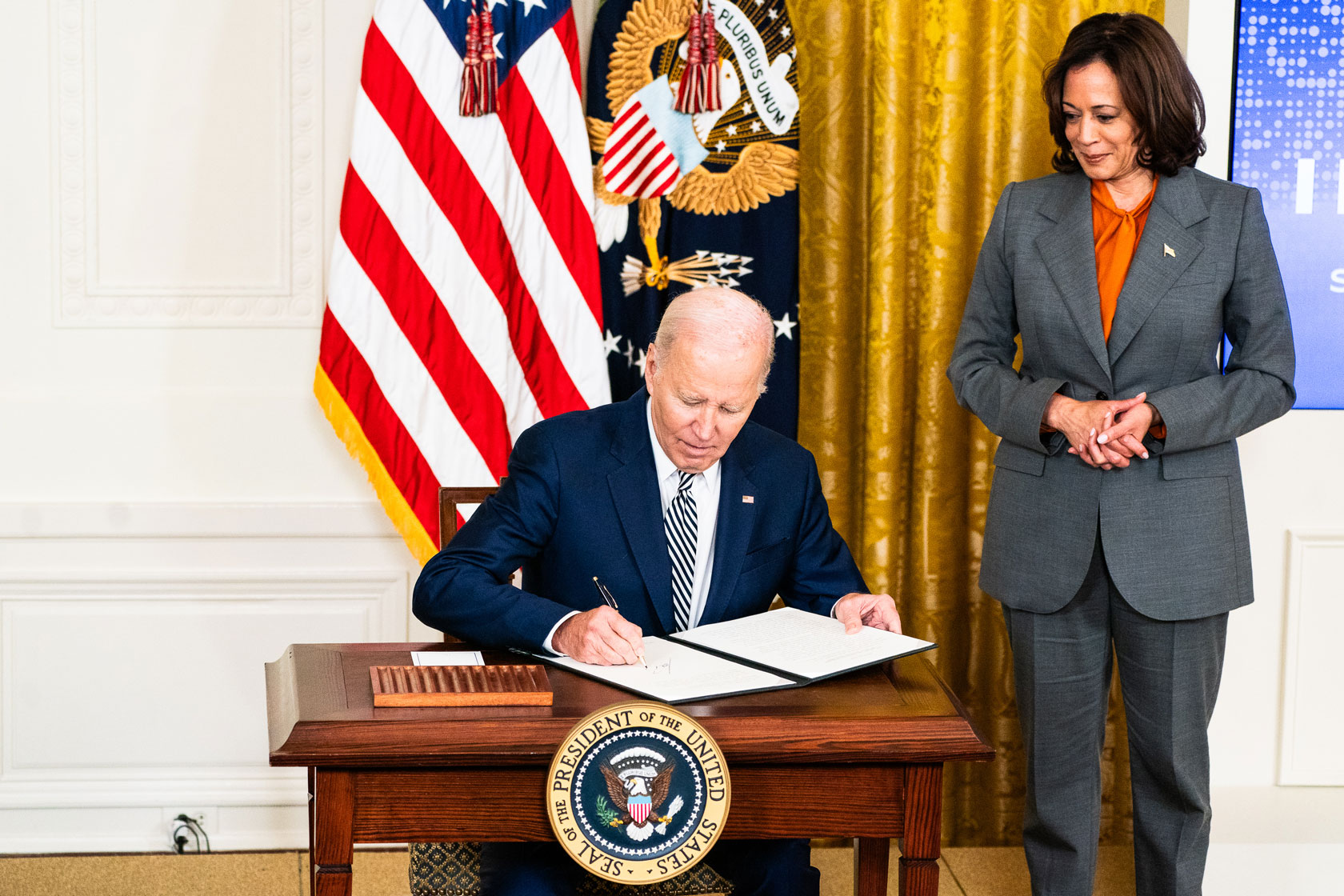 U.S. President Joe Biden sits at a desk with Vice President Kamala Harris standing to the right.